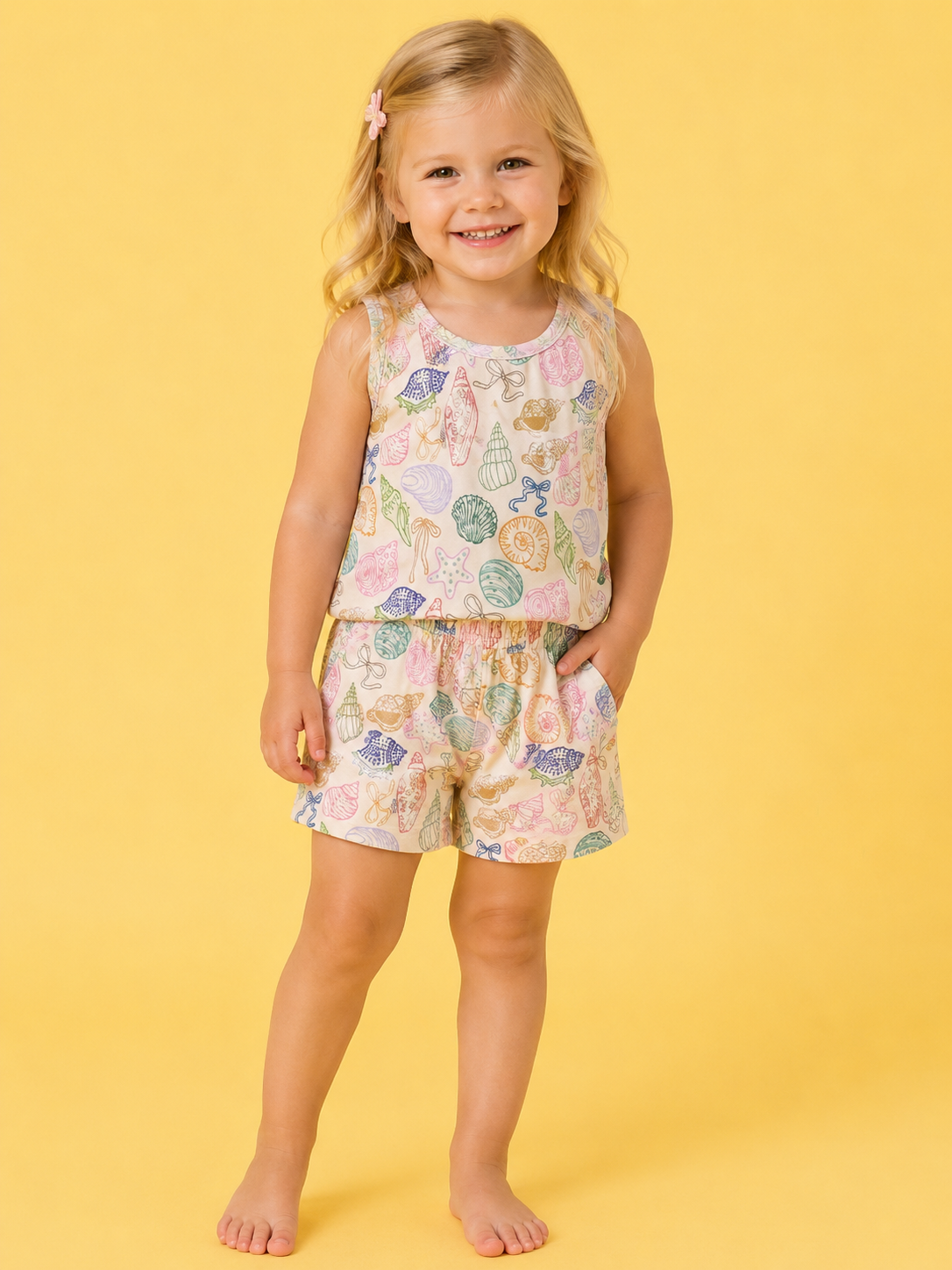 A smiling young girl stands barefoot against a yellow background, wearing the Treasure Island Tank Set by Tiny Tot Bundles—featuring a sleeveless top and shorts with a colorful seashell pattern.