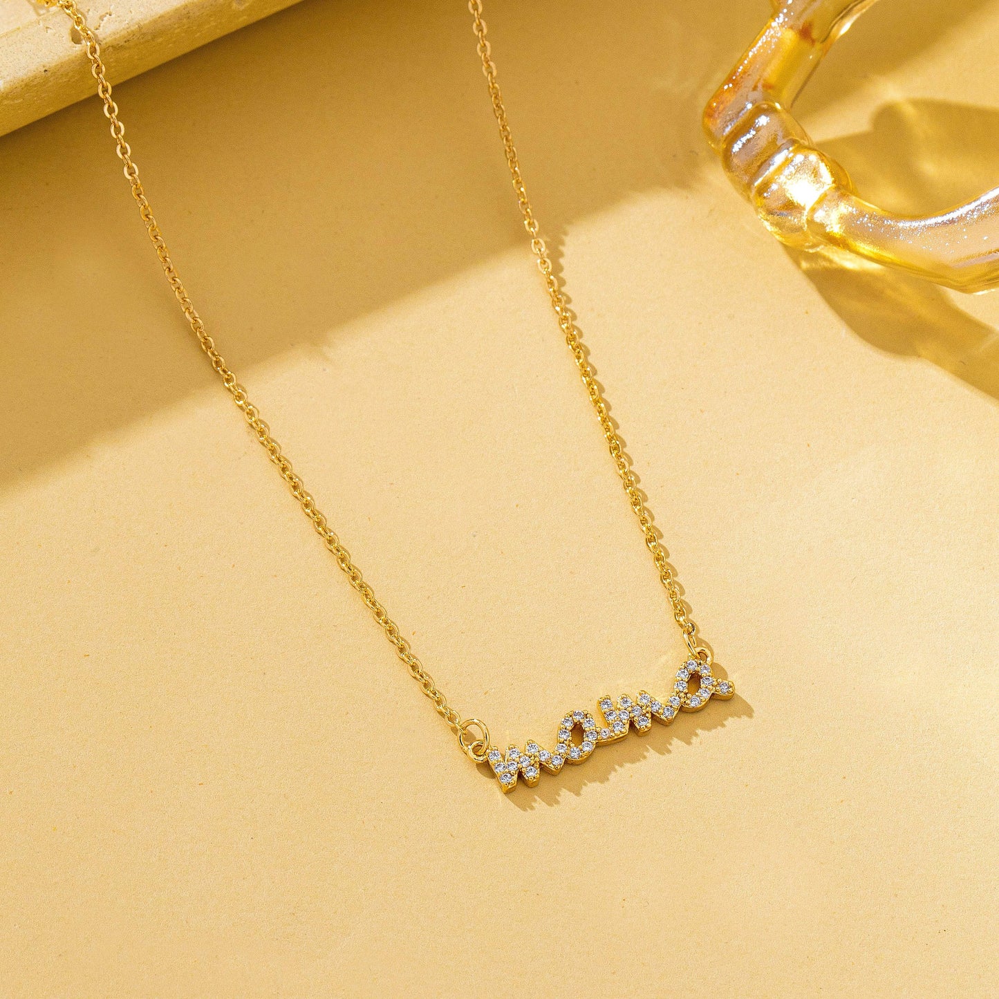 The JOSSLYN by wall to wall Gold MAMA Necklace, featuring a cursive pendant adorned with small sparkling stones, is displayed on a beige surface beside a transparent, light gold glass object.