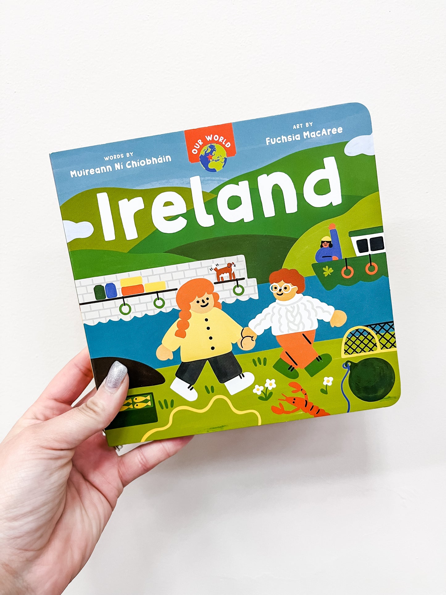 A hand holds Barefoot Books' "Our World: Ireland Book," featuring a vibrant cover with two smiling children, green hills, and Irish symbols.