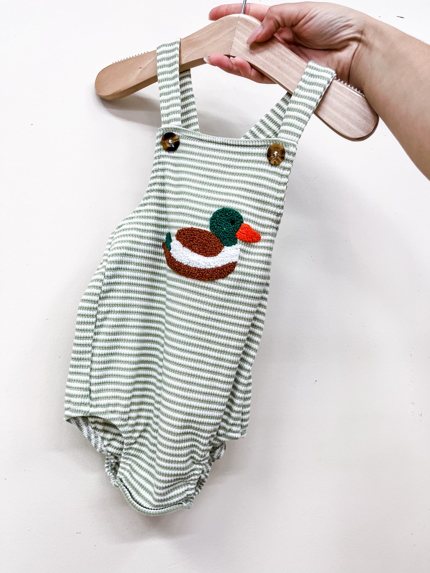 A hand displays the Tiny Tot Bundles Duck Stripe Romper—green and white stripes, wooden buttons, and a colorful embroidered duck—on a wooden hanger against a white background.