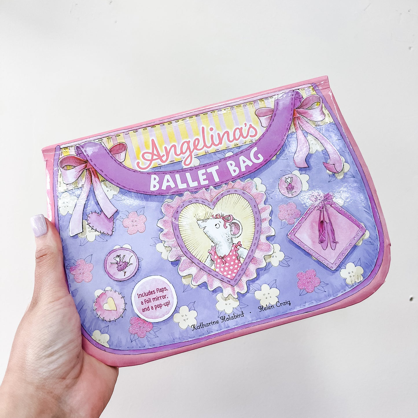 A hand holds Simon & Schuster's "Angelina's Ballet Bag Book," a purple and pink children's book featuring bows, ballet slippers, flowers, and a mouse in a tutu inside a heart-shaped frame.