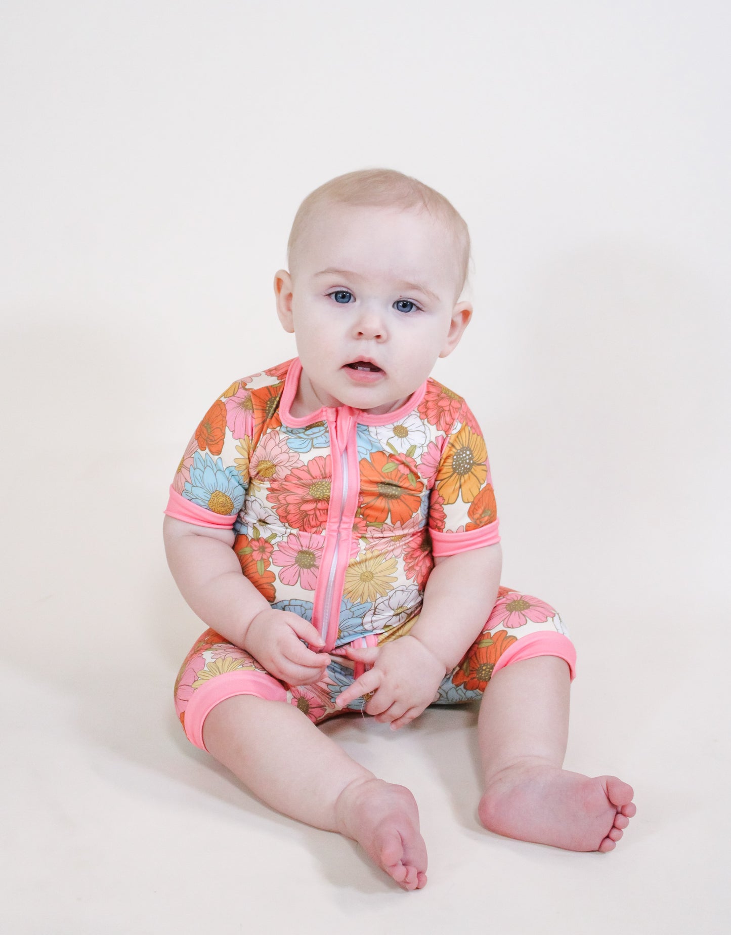 A light-skinned baby with short blonde hair wears Tiny Tot Bundles’ Retro Bloom Shorty, a colorful floral outfit with pink trim and zipper, while sitting on a light surface and looking slightly toward the camera.