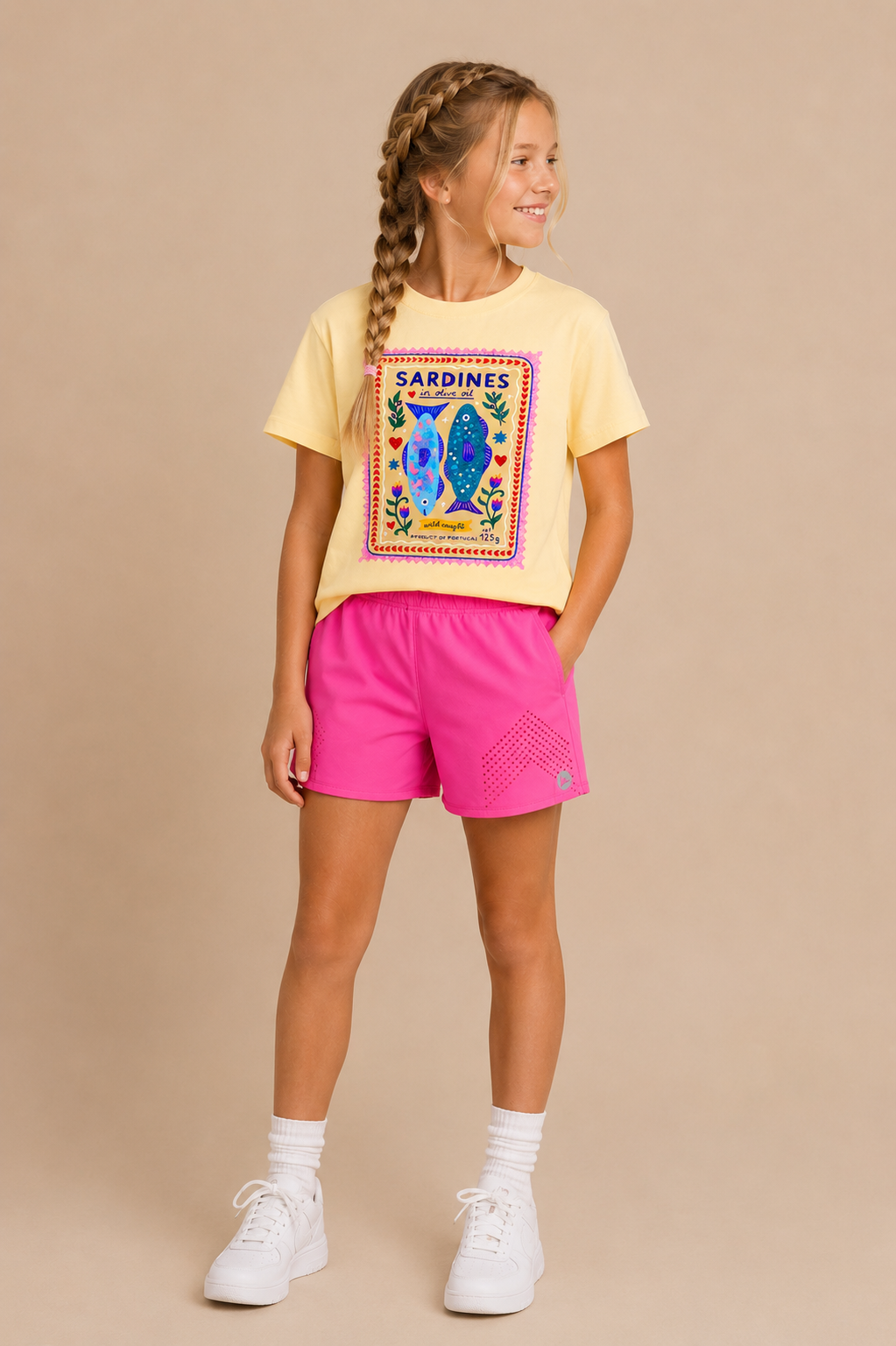 A young girl smiles and looks to the side, wearing the Sardines Youth Tee from Tiny Tot Bundles—featuring colorful sardine graphics—paired with bright pink shorts, white socks, and white sneakers against a beige background.