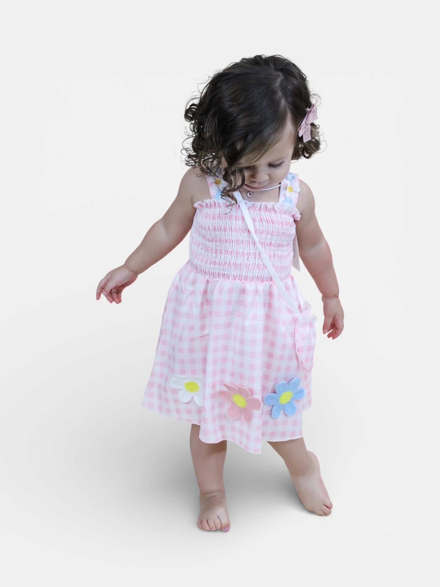A young child with curly hair stands barefoot against a white background, wearing Tiny Tot Bundles’ Petal Picnic Smocked Set—a light pink checkered outfit adorned with colorful fabric flowers and a matching crossbody bag.