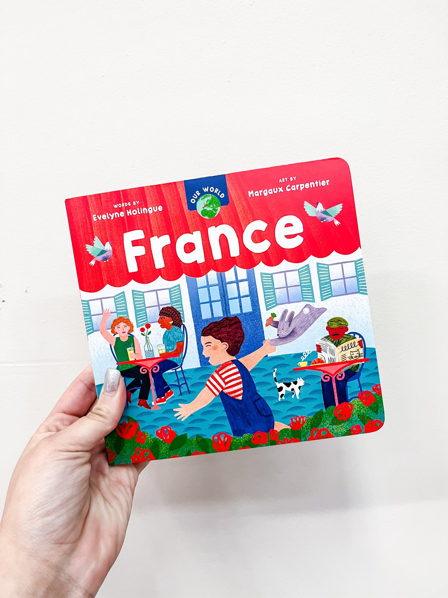 A hand holds Barefoot Books’ Our World: France Book, featuring vibrant illustrations of people at a French café, a child with a baguette, a black and white dog, and a market stand filled with cheese and fruit.