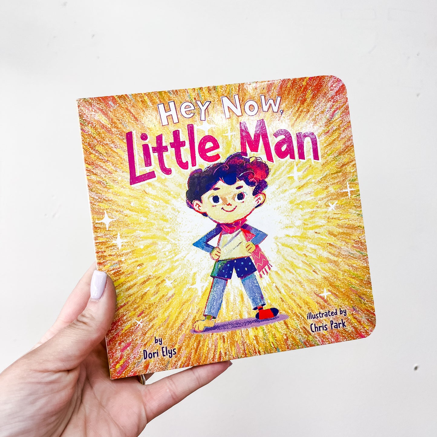 A hand holds the Simon & Schuster “Hey, Now, Little Man Book,” featuring a confident boy with hands on hips on a vibrant, colorful cover.