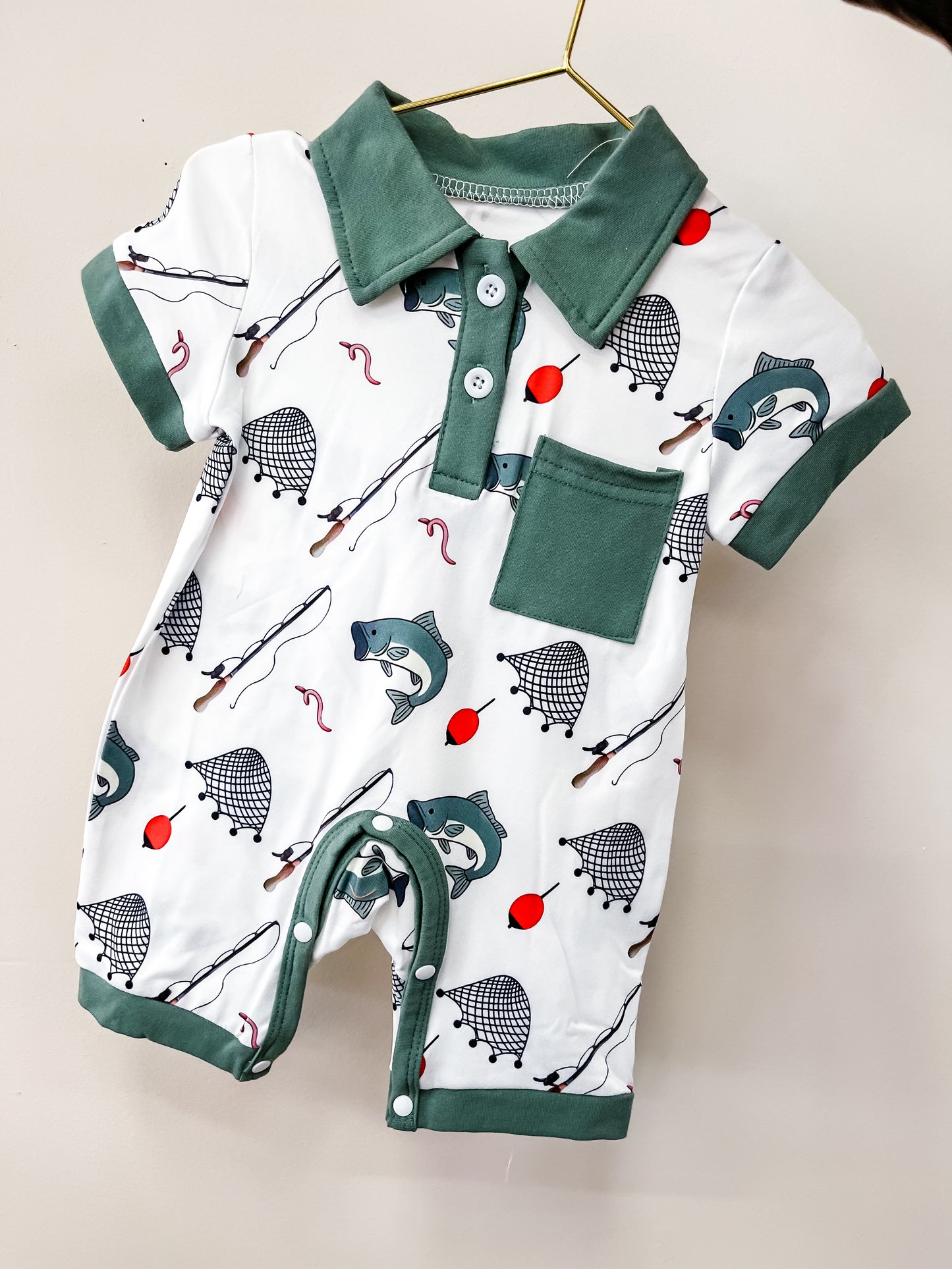 The Reel Cool Romper by Tiny Tot Bundles hangs on a gold hanger, featuring green trim, buttons, and a pocket with a fishing print—fish, nets, hooks, and bobbers—on a white background.
