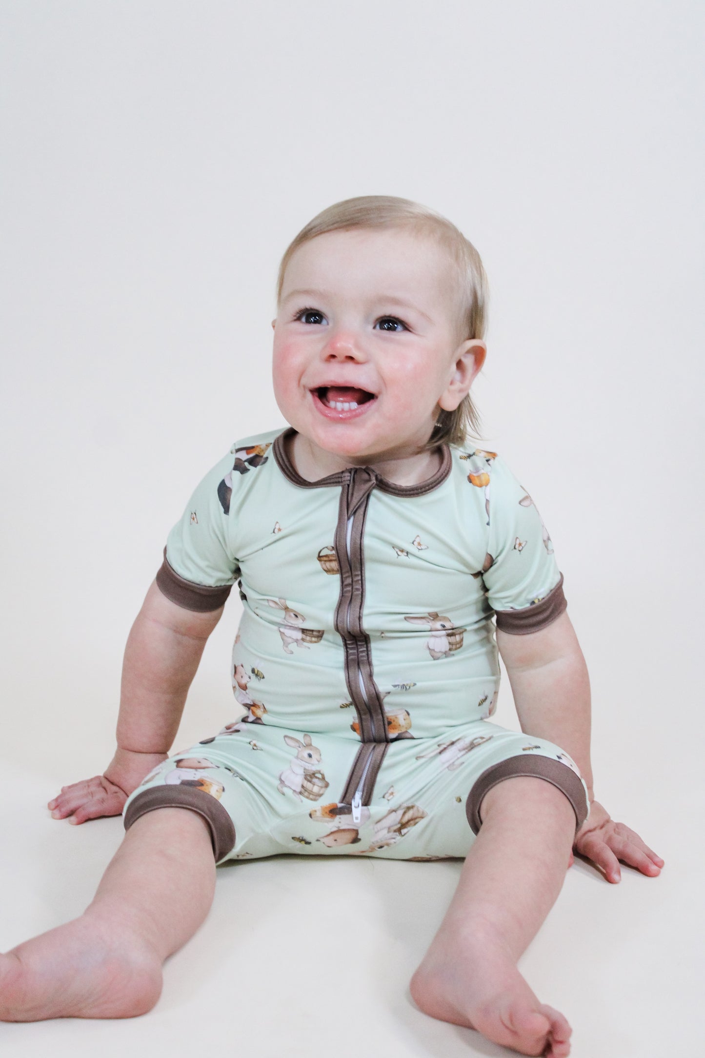 A smiling toddler with light hair sits barefoot on a light background, wearing the Tiny Tot Bundles Peter's Honey Adventure Shorty Zippy, a light green romper with short sleeves, brown zipper, and bear print.