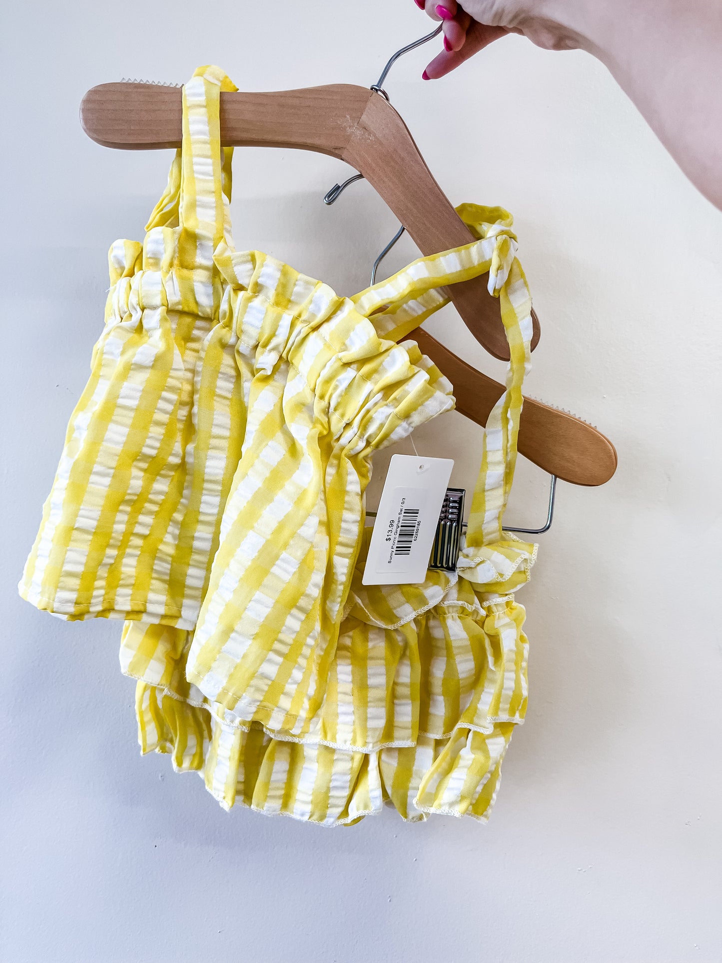 A hand holds a hanger with the Tiny Tot Bundles "Sunny Picnic Gingham Set," a yellow and white striped ruffled baby outfit with tags attached, shown against a plain white background.