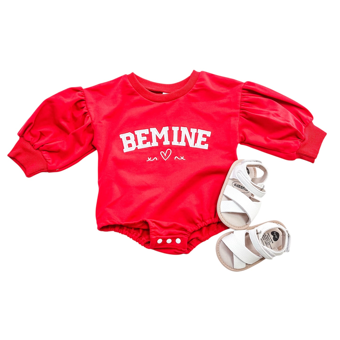 The Be Mine Puff Sleeve Bubble by Tiny Tot Bundles, a red baby romper with BE MINE and heart prints and puffy sleeves, is paired with white baby sandals on a white background.