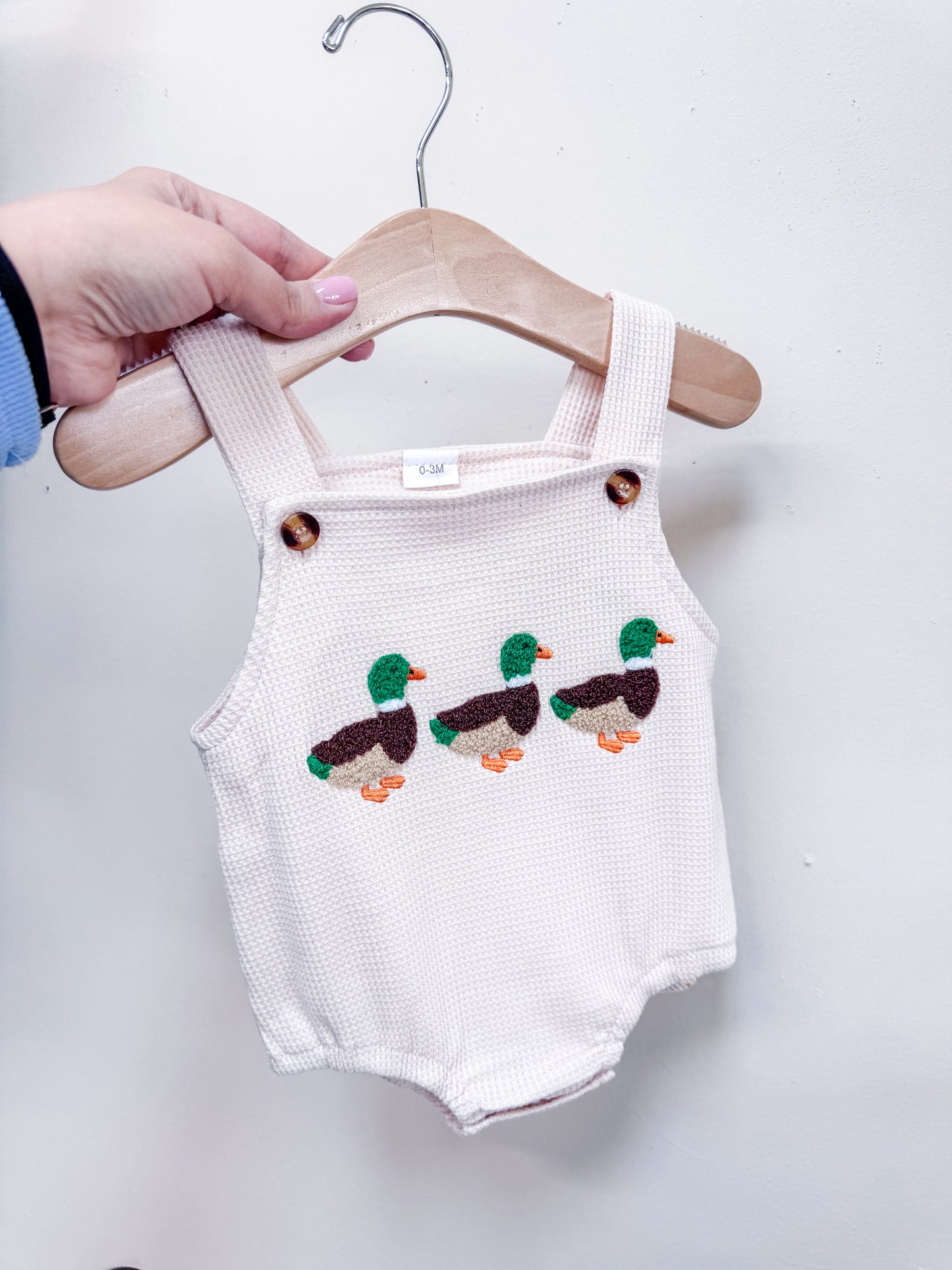 A hand holds Tiny Tot Bundles’ Little Duckling Romper, a white baby romper with straps and three embroidered mallard ducks on the front, displayed on a wooden hanger against a plain white wall.