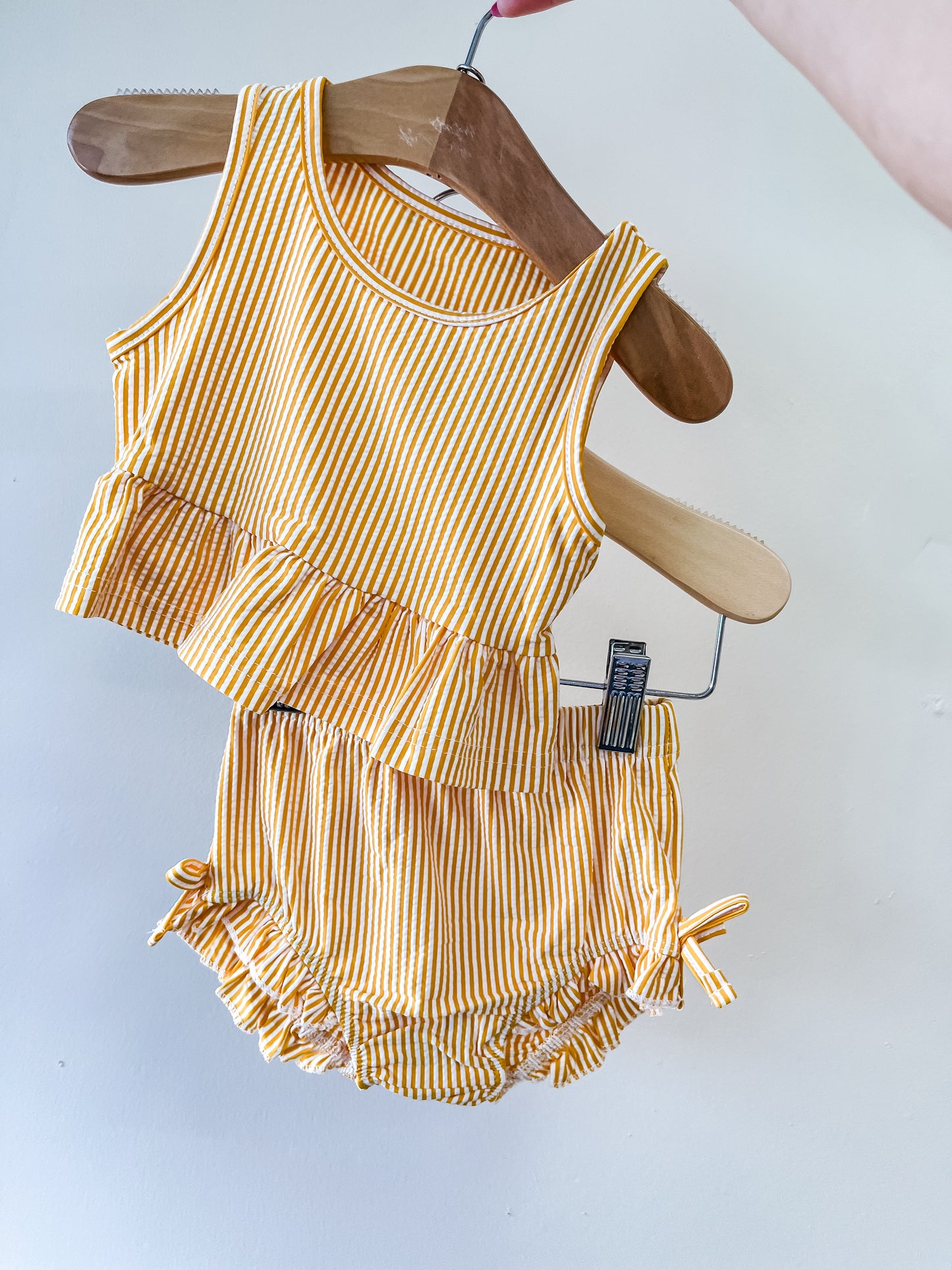 A hand holds a wooden hanger displaying the Tiny Tot Bundles Sunshine Stripe Swim Set—a yellow and white striped baby outfit with a sleeveless ruffled top and matching bloomers featuring small side bows.