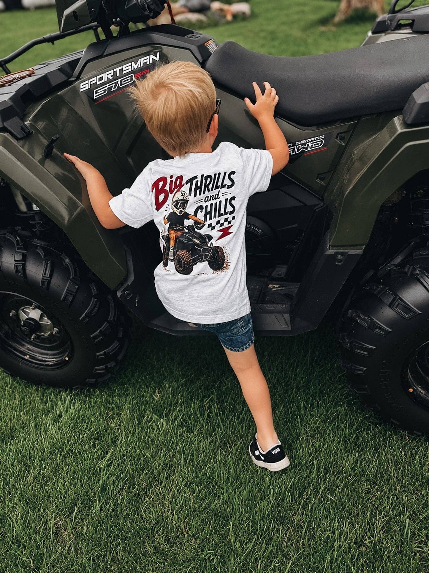 A young child wearing a Raising Brave Big Thrills and Chills Toddler Tee stands on the grass beside a dark green ATV, reaching up to touch the seat.