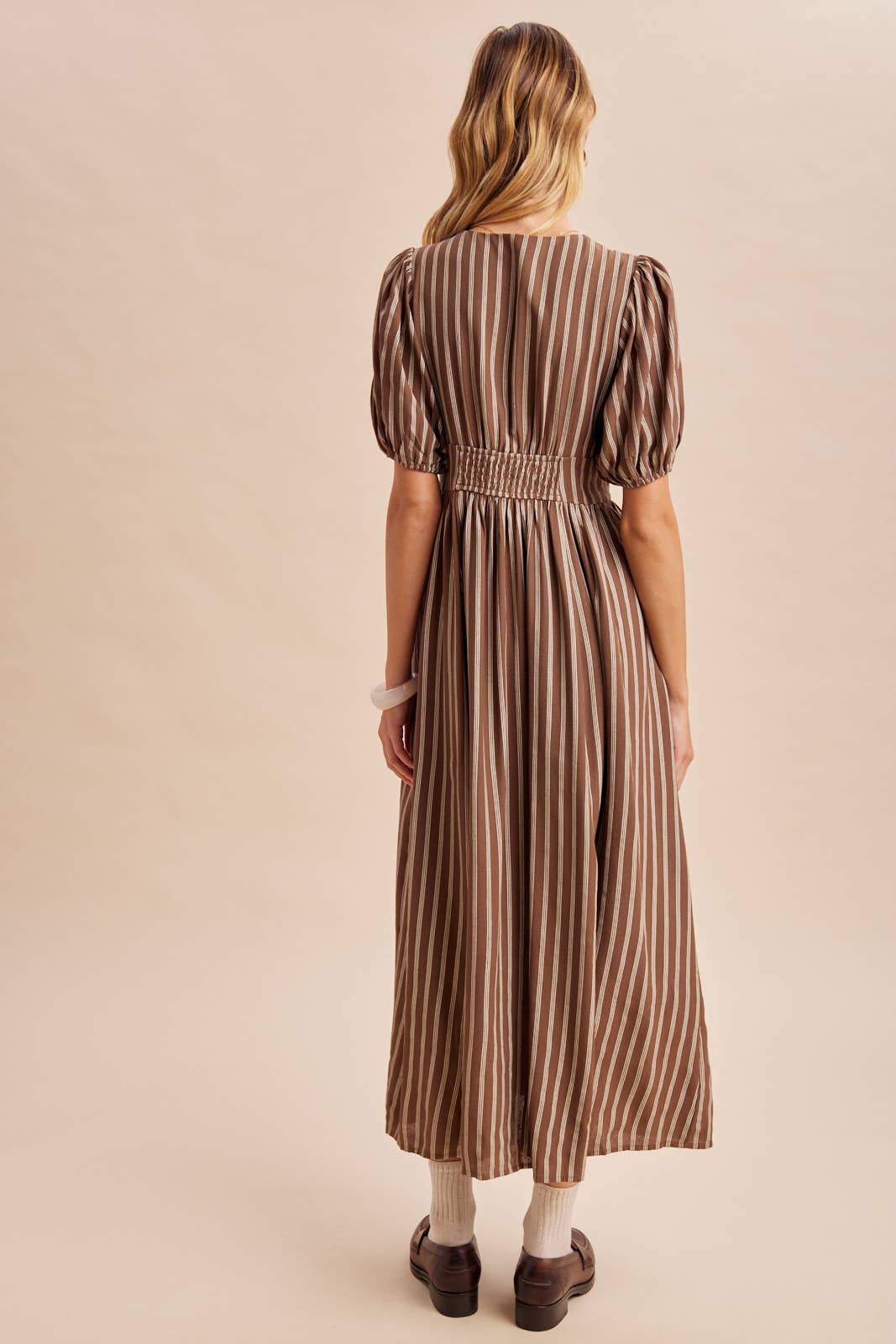 A person seen from behind wears an In Loom Stripe Button Down Maxi Dress, featuring brown and white vertical stripes, puffed sleeves, a gathered waist, beige socks, and dark loafers.