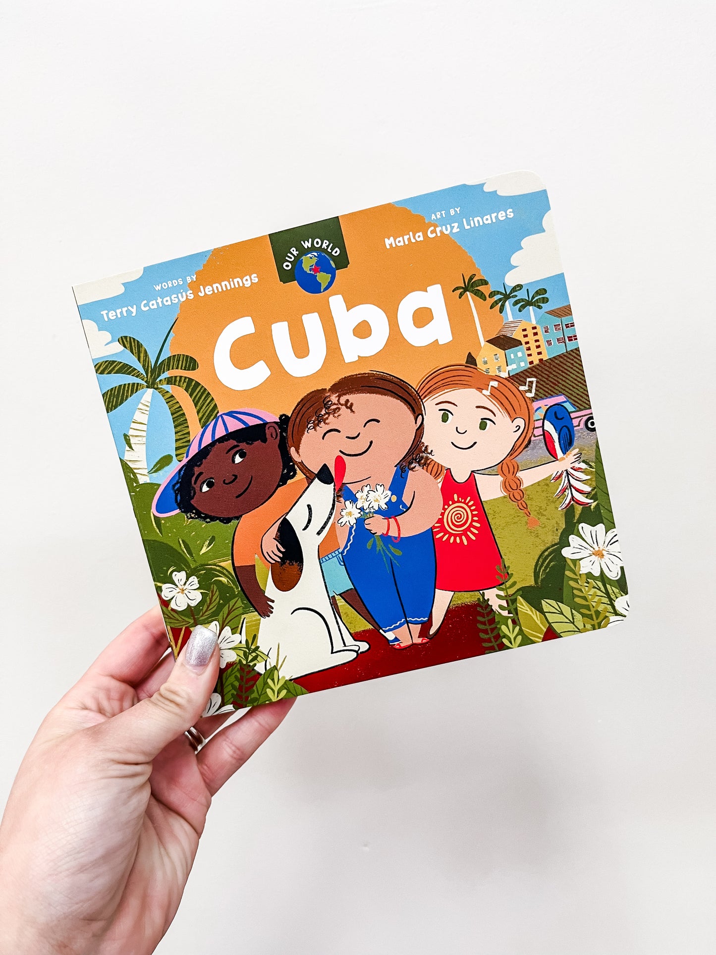 A hand holds Barefoot Books' "Our World: Cuba Book," featuring an illustrated cover with three smiling children, palm trees, and buildings, creating a cheerful tropical vibe.