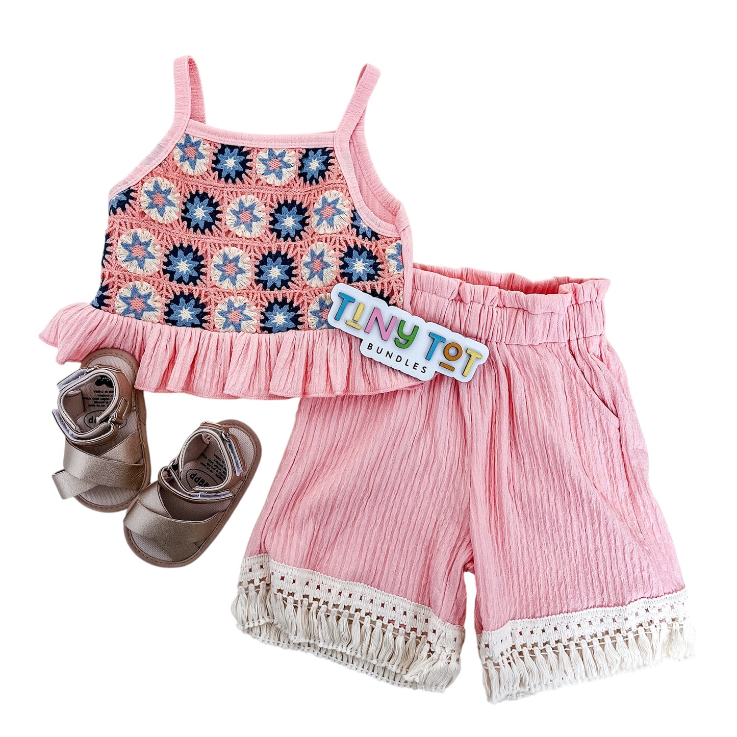 The Liliana Two Piece Set by Tiny Tot Bundles features a pink crochet tank top with blue and white accents, matching fringe-trimmed pink shorts, coordinating light brown sandals, and comes with a branded tag.