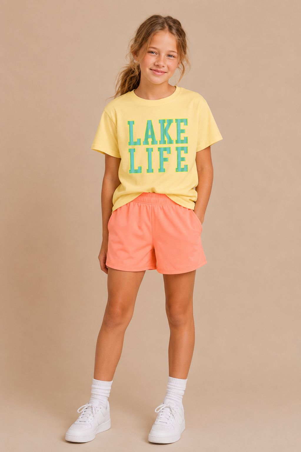 A young girl smiles in a Tiny Tot Bundles Lake Life Youth Tee, featuring green and blue lettering, paired with peach shorts, white crew socks, and white sneakers against a neutral beige background.