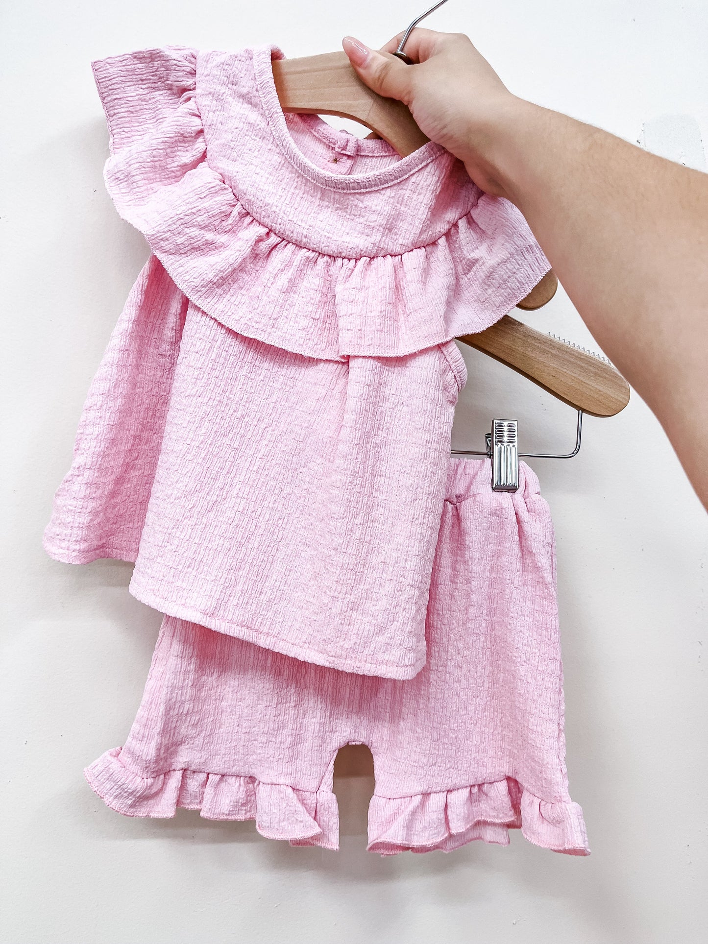 A hand holds a wooden hanger with the Pink Flutter Summer Set by Tiny Tot Bundles—a pink textured baby outfit featuring a ruffled top and matching ruffled shorts—against a plain white background.