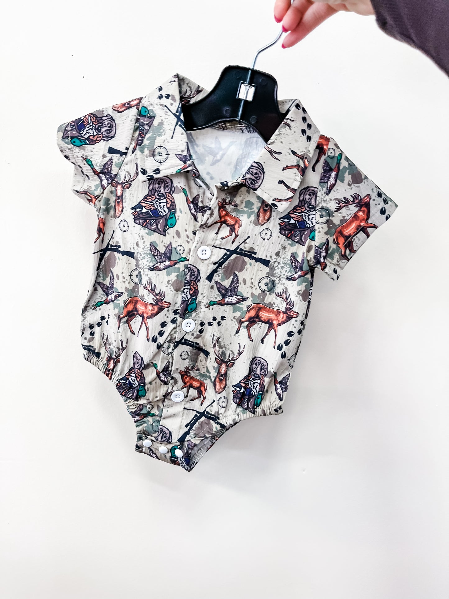 A hand holds a hanger displaying the Little Hunter Bubble by Tiny Tot Bundles—a short-sleeve baby onesie with woodland animal prints, buttons down the front, and a collar on a white background.