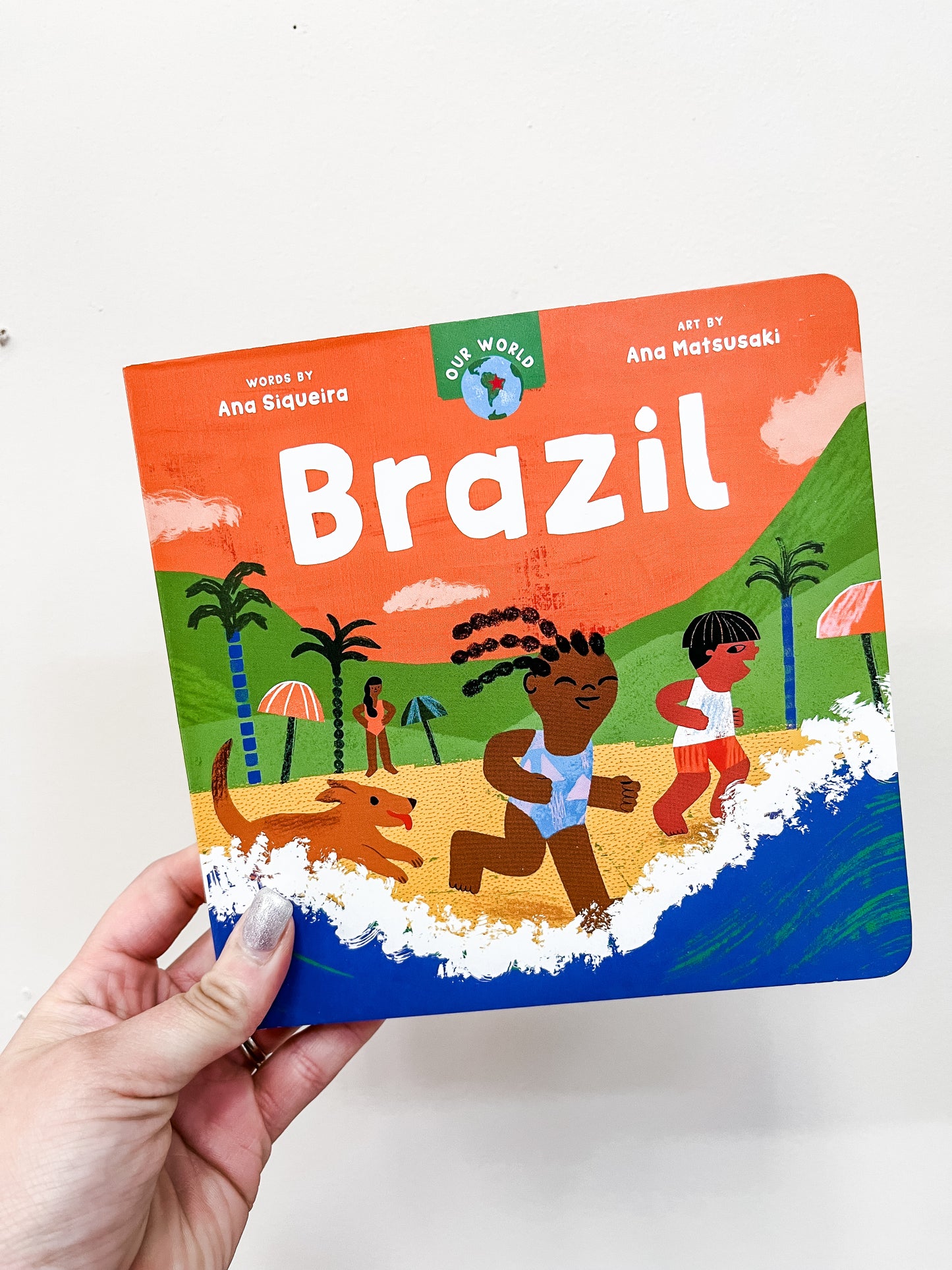 A hand holds Barefoot Books' "Our World: Brazil Book," featuring illustrations of two children and a dog playing on a beach with palm trees and ocean waves in the background.
