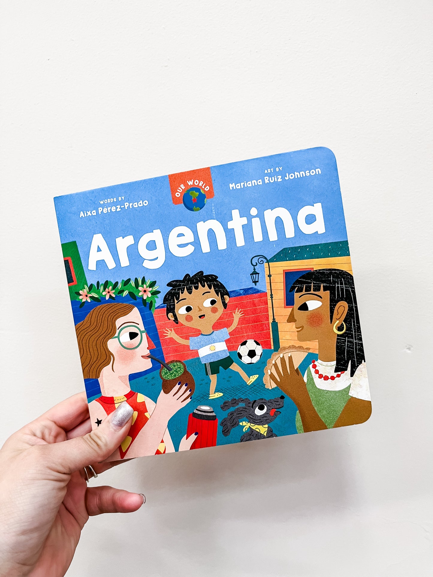 A hand holds Barefoot Books’ “Our World: Argentina Book,” a children’s book with a colorful cover showing cartoon people, a dog, soccer ball, and traditional drink.