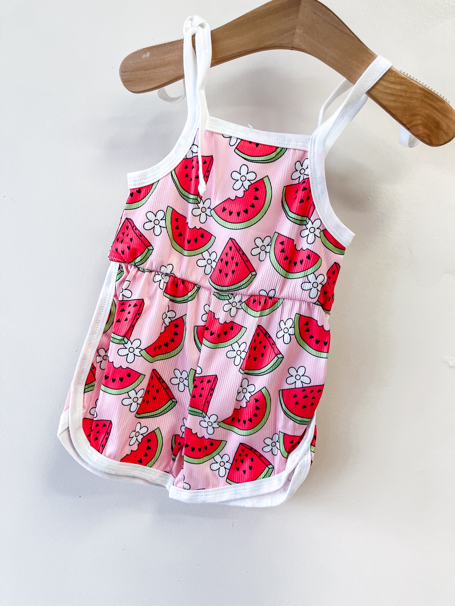 The Sweet Watermelon Romper by Tiny Tot Bundles is a pink toddler outfit with white straps and trim, featuring playful watermelon and white flower prints, displayed on a wooden hanger against a white wall.