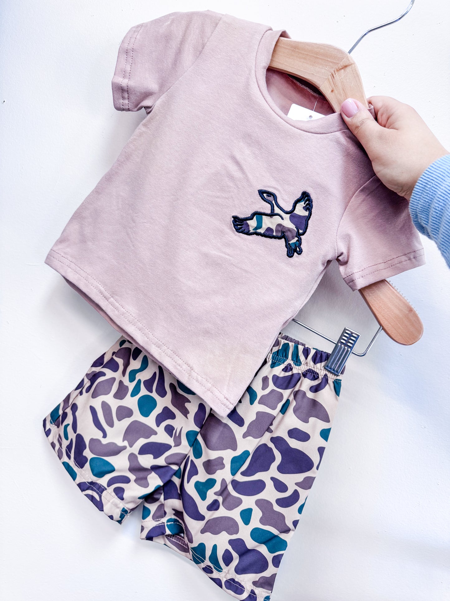 A hand displays the Little Hunt Duo Boys Set by Tiny Tot Bundles—a light pink short-sleeve shirt with a camouflage bunny patch, paired with matching purple, green, and beige camouflage shorts.