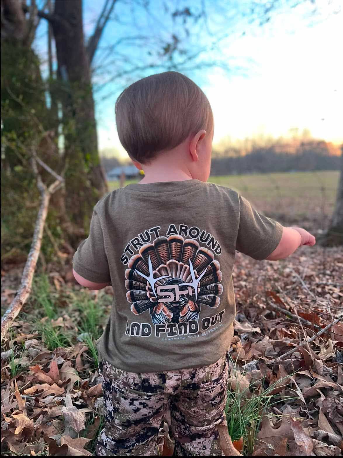 A toddler seen from behind wearing a brown Southern Hyde Co. Strut Around and Find Out Tee featuring a turkey graphic.