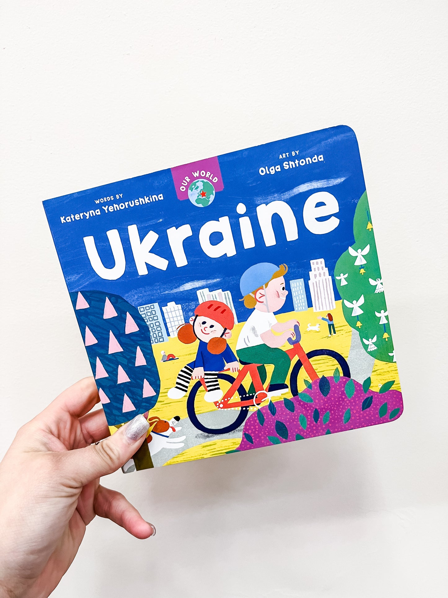A hand holds Barefoot Books’ "Our World: Ukraine," a colorful children's book featuring an illustration of two kids riding a bicycle past city buildings and greenery.
