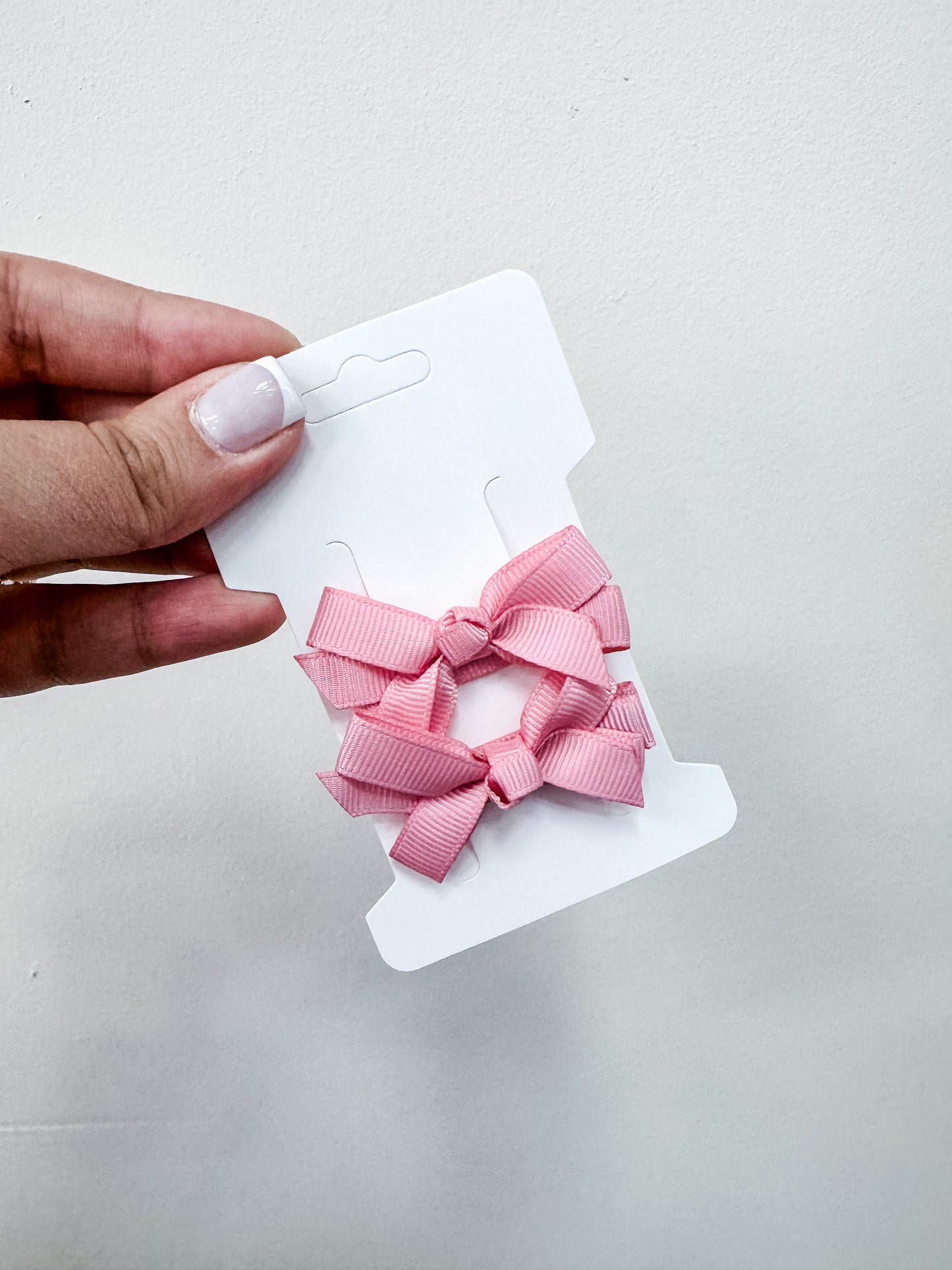 A hand with manicured nails holds a white card displaying Tiny Tot Bundles' Duo Piggy Clip Pack—two small pink ribbon hair bows—against a plain, light background.