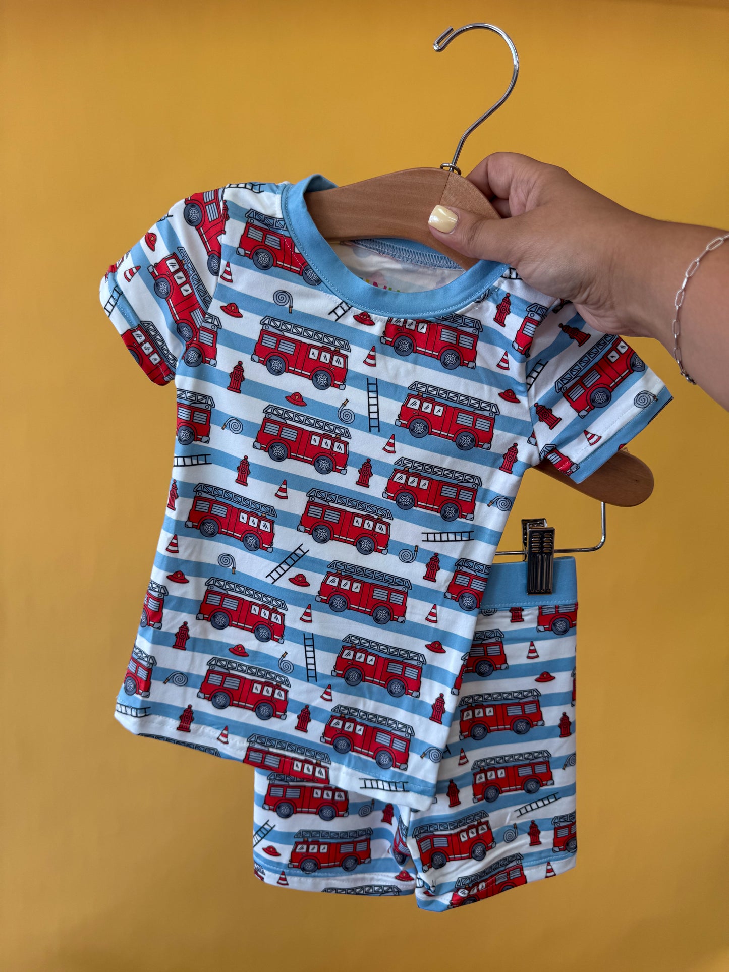 A hand holds a hanger displaying the Tiny Tot Bundles Rescue Ready Lounge Set, a matching children’s shirt and shorts ensemble adorned with a fire truck pattern, set against a solid yellow background.
