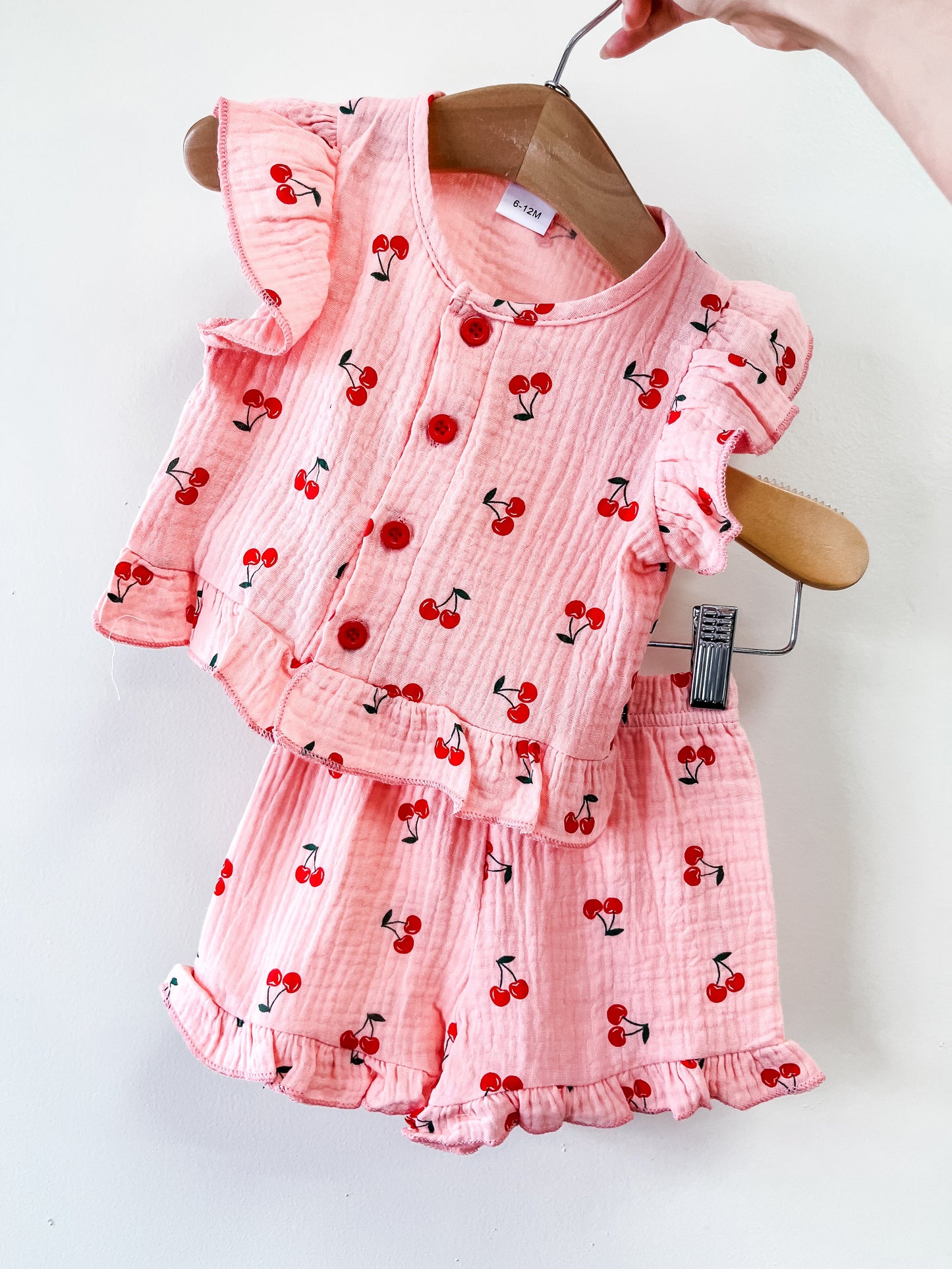 The Linen Chery Pink Set by Tiny Tot Bundles, featuring a pink ruffled cherry-print top and matching shorts, is displayed on a wooden hanger against a white background.