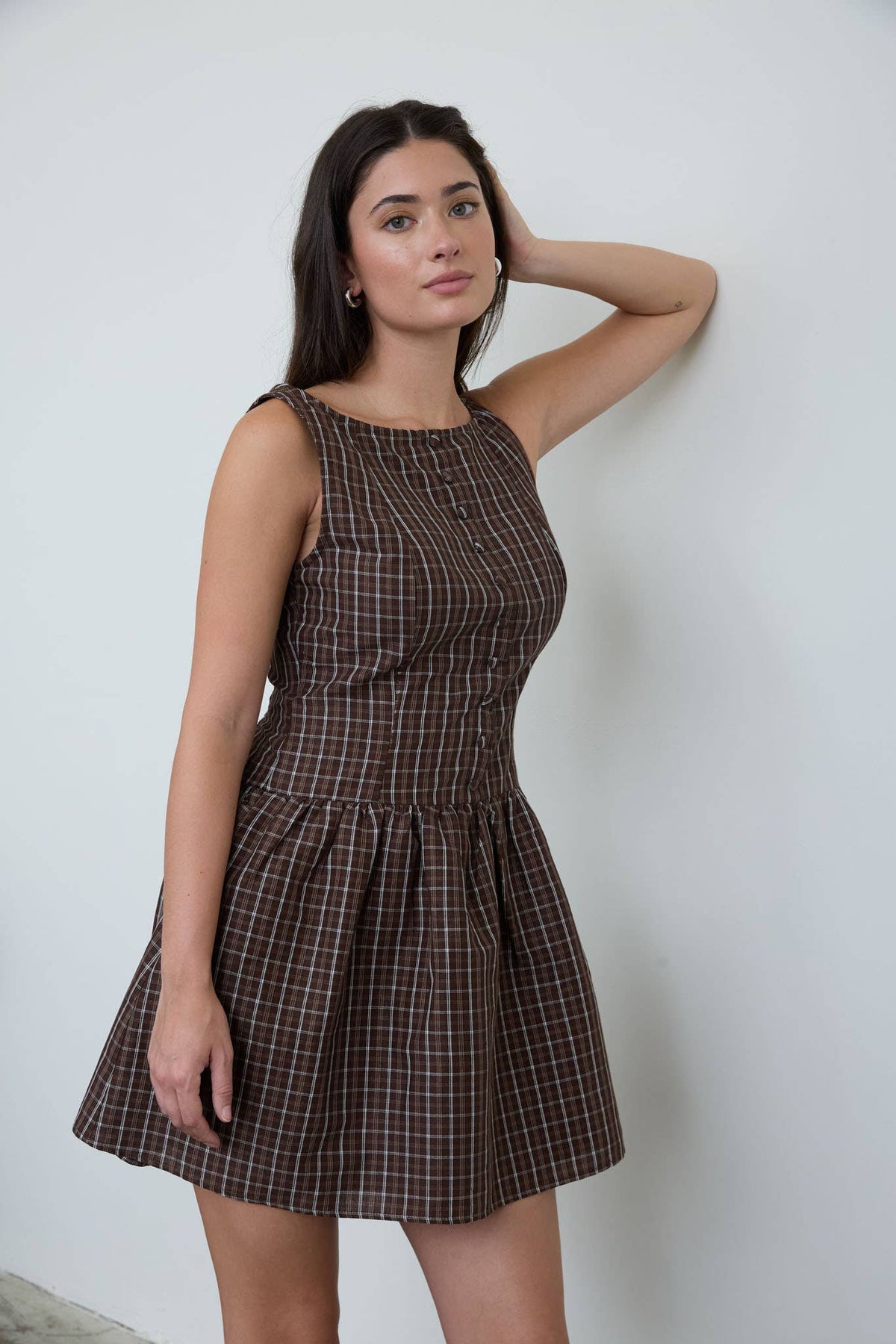 Wearing The Workshop Studio’s Brown Plaid Boat Neck Dress, a woman stands against a white wall, one arm bent with her hand touching her head, gazing at the camera.
