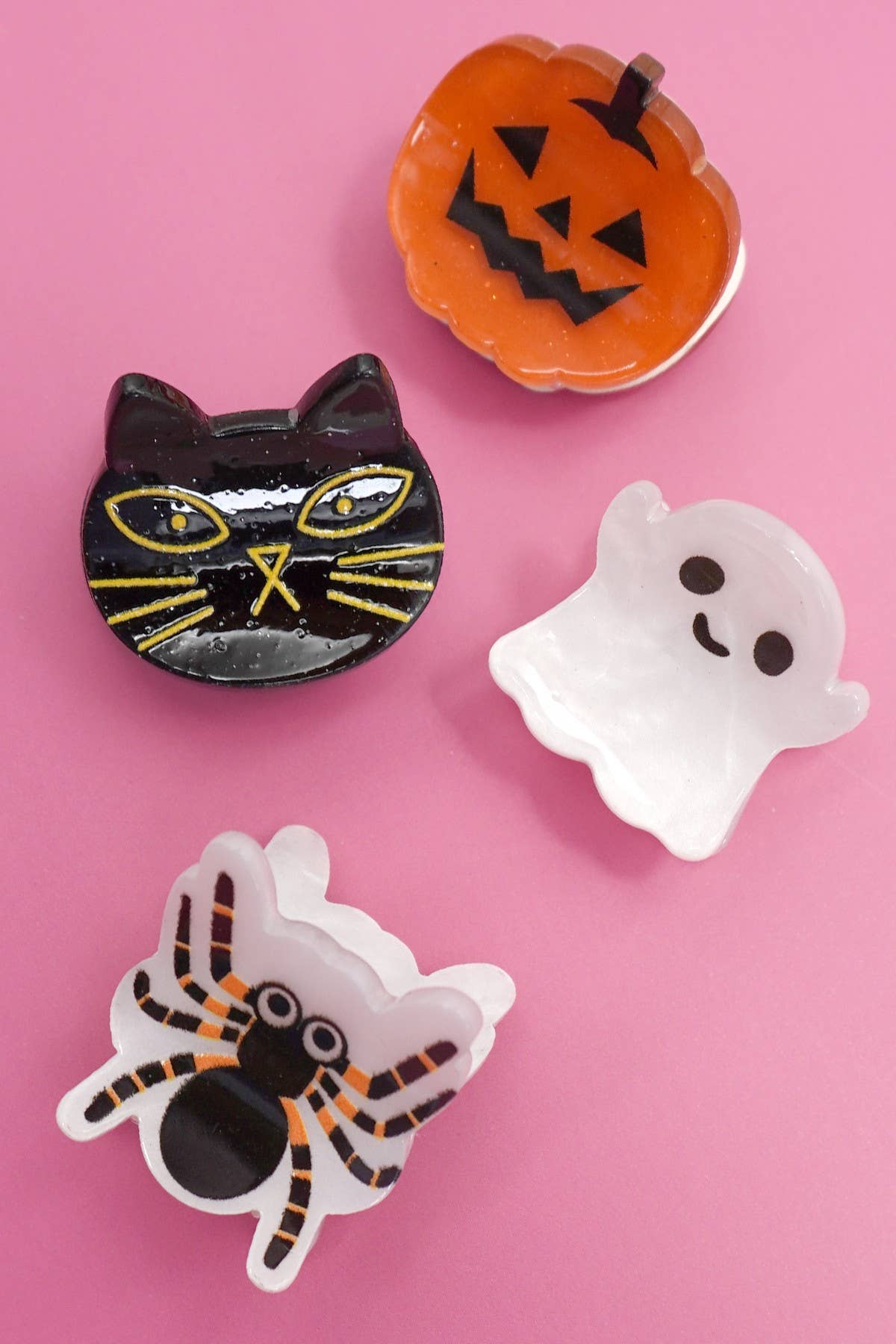 Four Halloween Ghost Theme Mini Hair Clips by JOSSLYN by wall to wall displayed on a pink background: an orange smiling pumpkin, a black cat with yellow eyes, a happy white ghost, and a black spider with white outline and orange details. Set includes 4 clips.