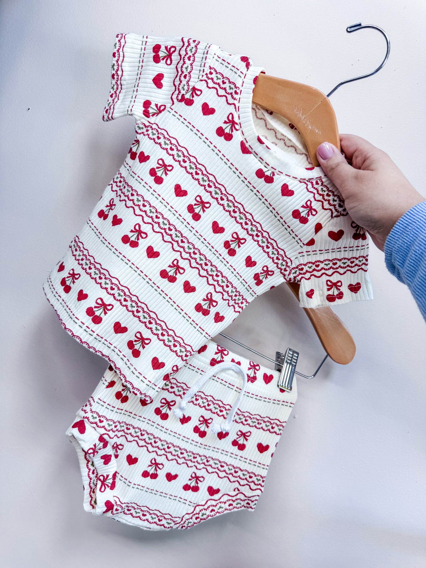 A hand holds the Cream Cherry Ribbed Set by Tiny Tot Bundles, a matching short-sleeve top and shorts featuring red cherry and bow patterns on white ribbed fabric, displayed on a hanger against a light-colored wall.