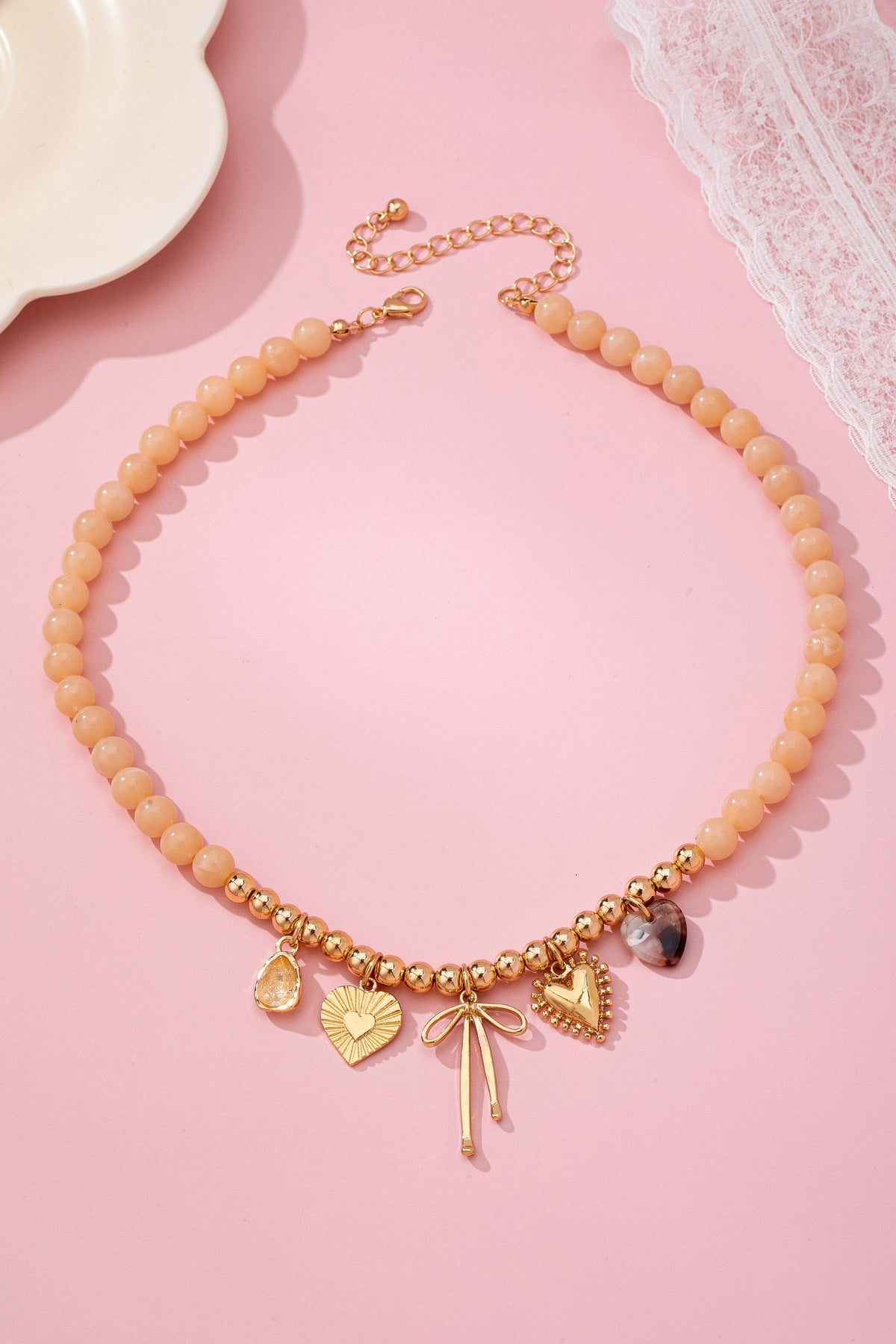 A Beige Beaded Bow Heart Rhinestone Charm Necklace by JOSSLYN by wall to wall, featuring gold charms—including a heart, leaf, dragonfly, and additional hearts—displayed on a pink surface accented with lace and a white dish in the corners.