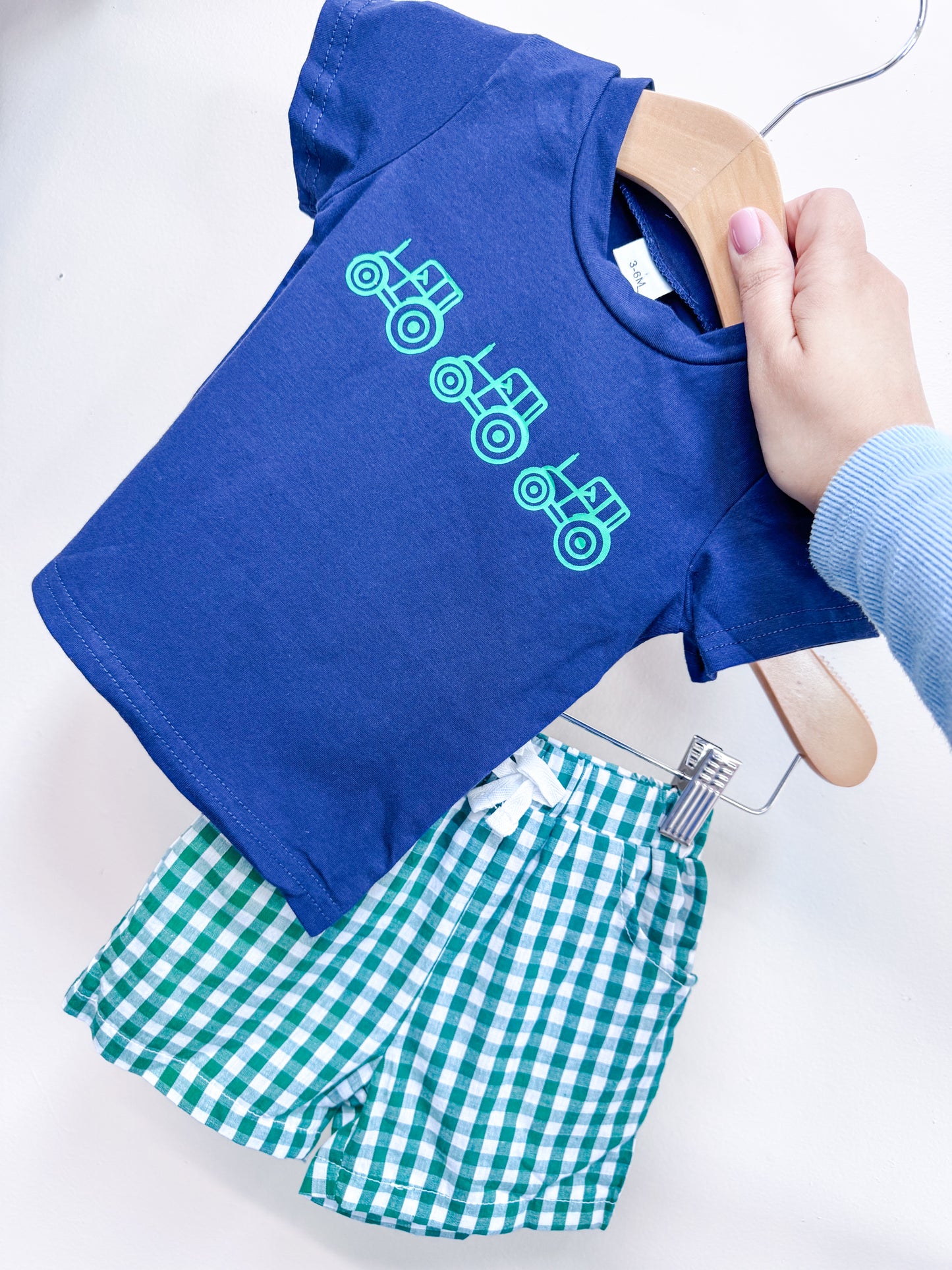 A hand holds the Little Farmer Boys Set by Tiny Tot Bundles, featuring a blue tee with three green tractor graphics and green-and-white checkered shorts, displayed against a plain light background.