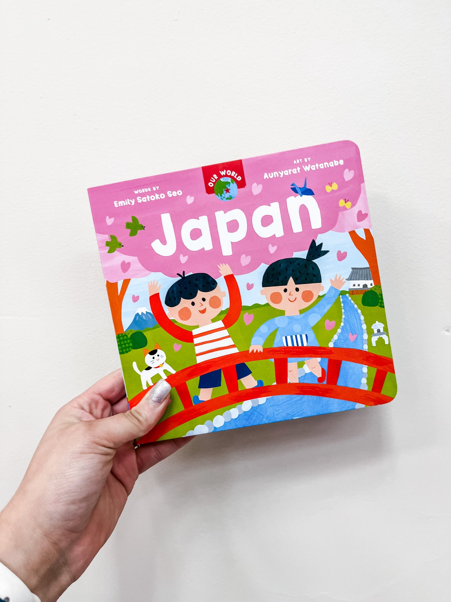 A hand holds Barefoot Books' Our World: Japan, a colorful children’s book showing two cartoon kids waving on a bridge with cherry blossoms, Mount Fuji, a dog, and a river in the background.