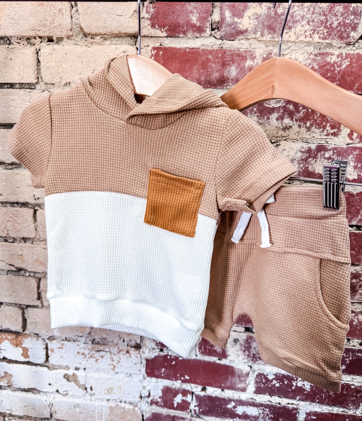 The Colorblock Tan Boys Set by Tiny Tot Bundles features a short-sleeve hooded top with tan, white, and a brown chest pocket plus matching tan waffle-knit shorts, shown hanging on wooden hangers against a brick wall.