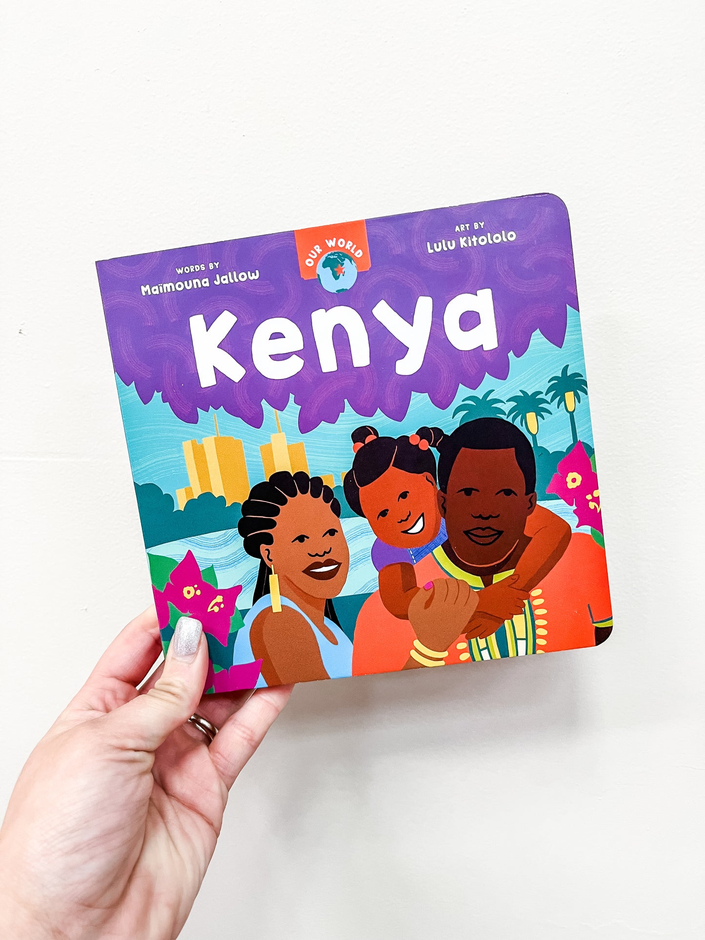A hand holds Barefoot Books’ "Our World: Kenya Book," featuring an illustrated, smiling family of three on the cover with a vibrant tropical backdrop of palm trees and buildings.