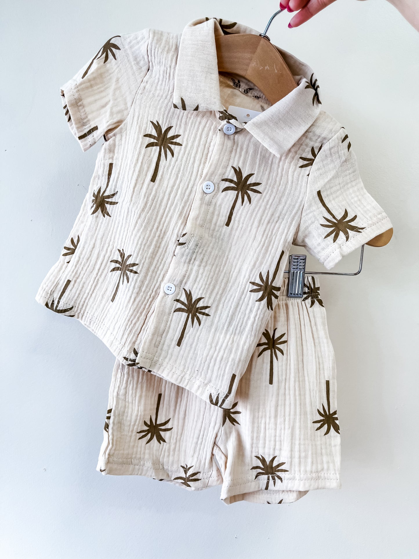 The Tiny Tot Bundles Palm Breeze Button Set, a beige baby outfit with brown palm tree prints, includes a short-sleeve button-up shirt and matching shorts, displayed on a hanger against a white background.