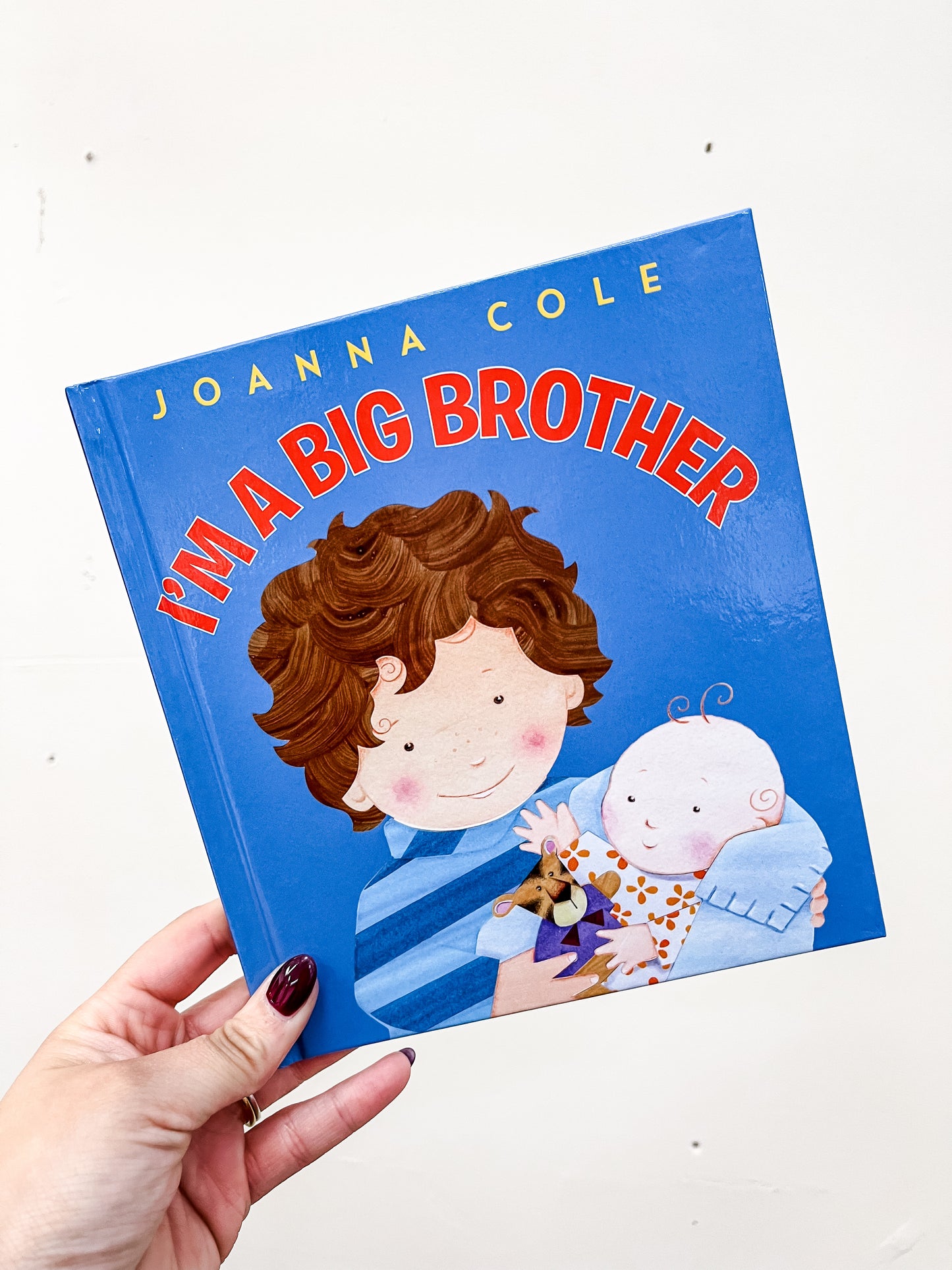 A hand holds the harper Collins "I'm A Big Brother Book," showing a smiling boy with a baby and stuffed animal on a blue background.