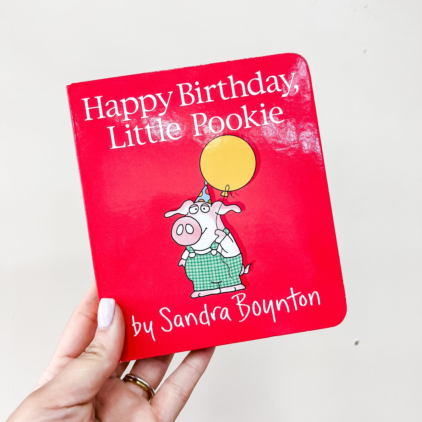 A hand holds the Happy Birthday, Little Pookie Book by Simon & Schuster, showing an illustrated pig in green overalls with a yellow balloon on the cover.