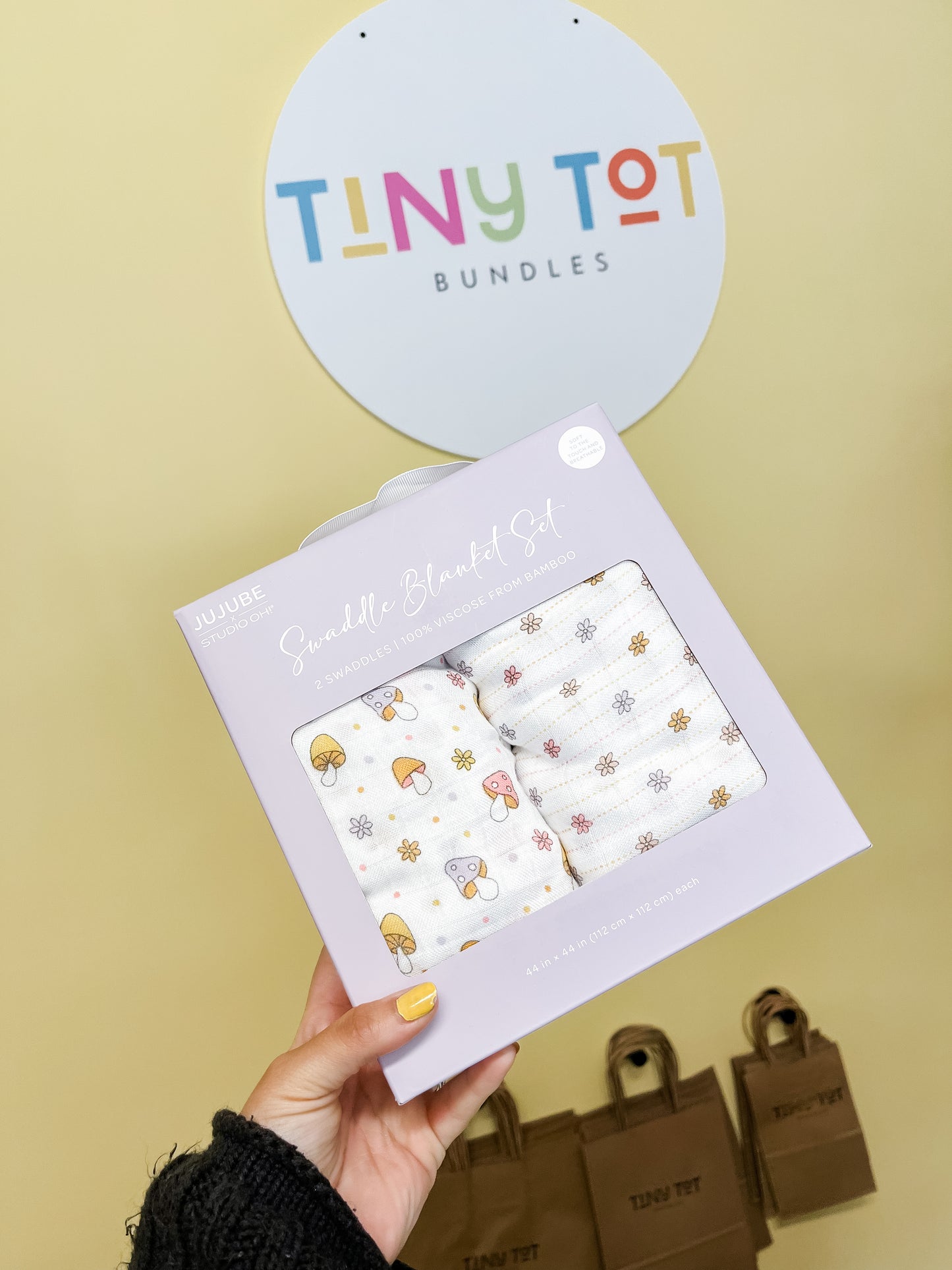 A hand holds a pastel purple Swaddle Blanket Set - Mushy Love from Tiny Tot Bundles. Behind it, a round Tiny Tot Bundles sign hangs on a yellow wall with brown gift bags on the floor.