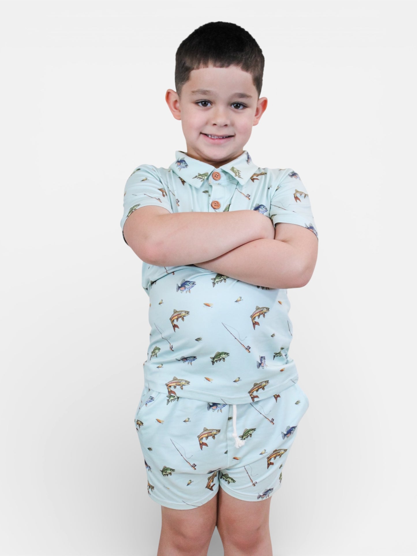 A young boy with dark hair smiles and stands with arms crossed, wearing the Tiny Tot Bundles Fishing Day Lounge Set—matching light-colored shirt and shorts featuring a fun print—against a plain white background.
