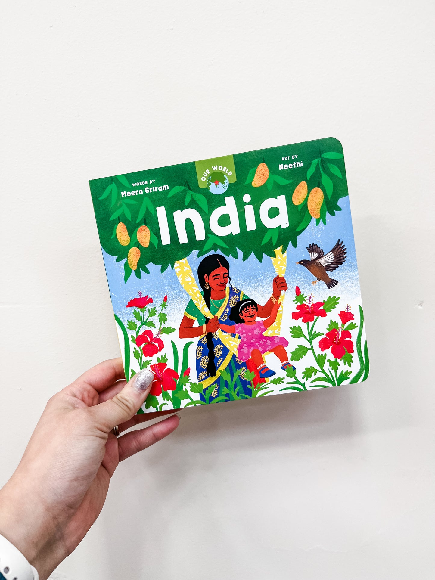 A hand holds Barefoot Books' "Our World: India," a colorful children's book featuring cover art by Neethi of a woman and child standing amid flowers, trees, mangoes, and a bird.