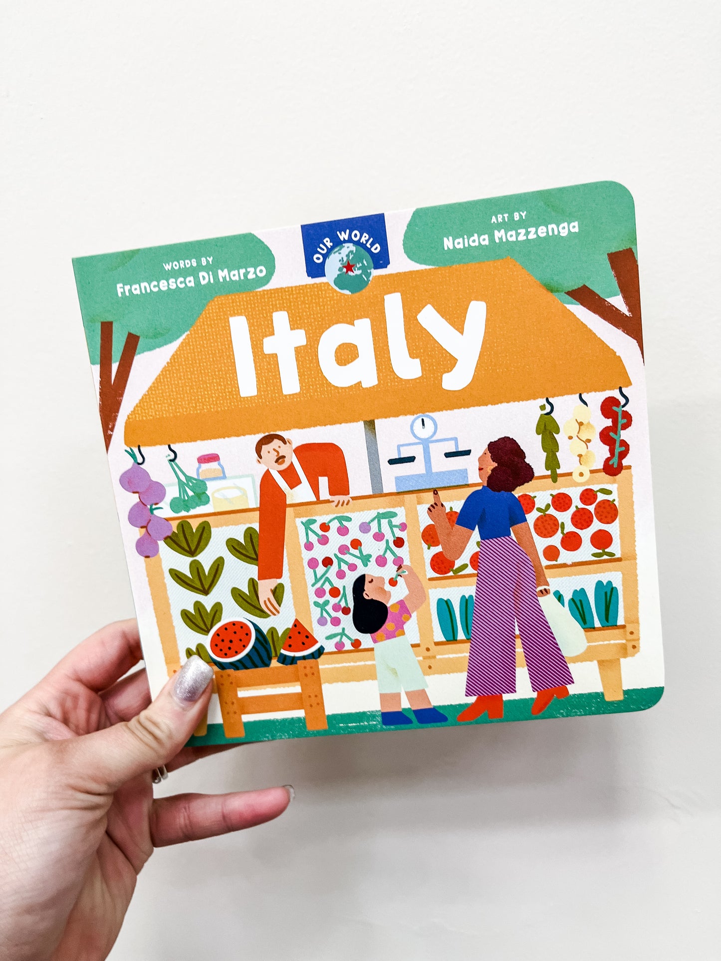 A hand holds Barefoot Books' "Our World: Italy Book," showing a vibrant cover with a woman, child, and vendor at a fruit and vegetable market. The cover credits Francesca Di Marzo and Naida Mazzenga.