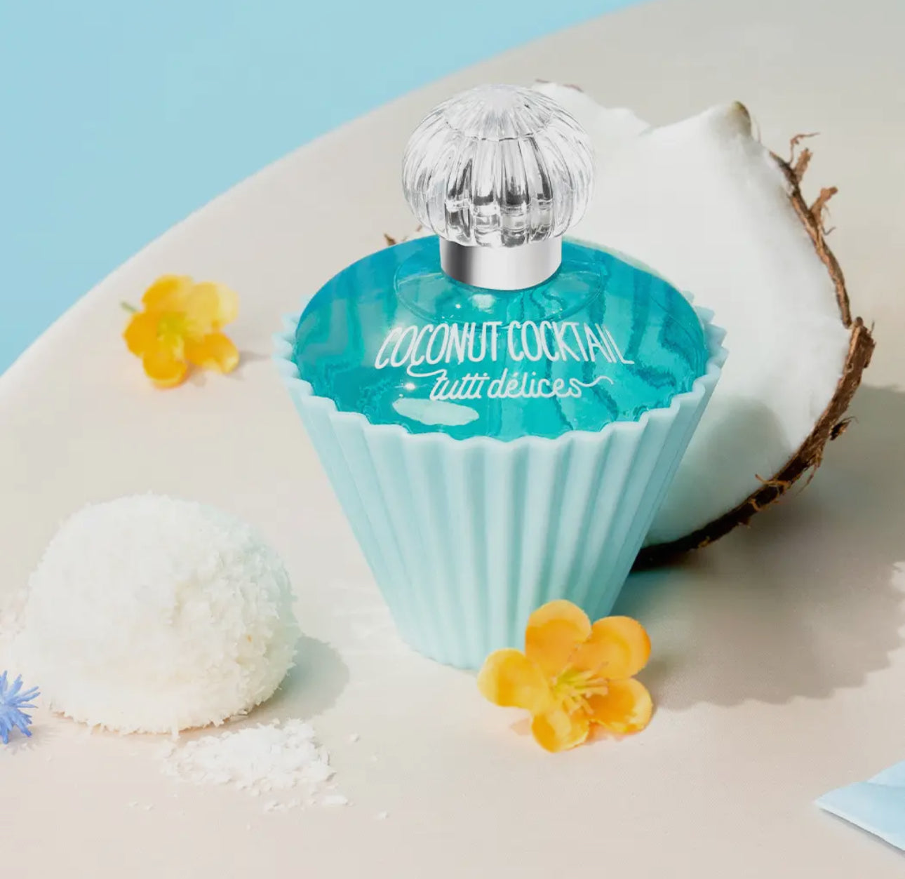 A turquoise, cupcake-shaped bottle labeled Tutti Delices Coconut Cocktail Cupcake Perfume sits next to a cracked coconut, white coconut ball, and yellow flowers on beige with a light blue background.