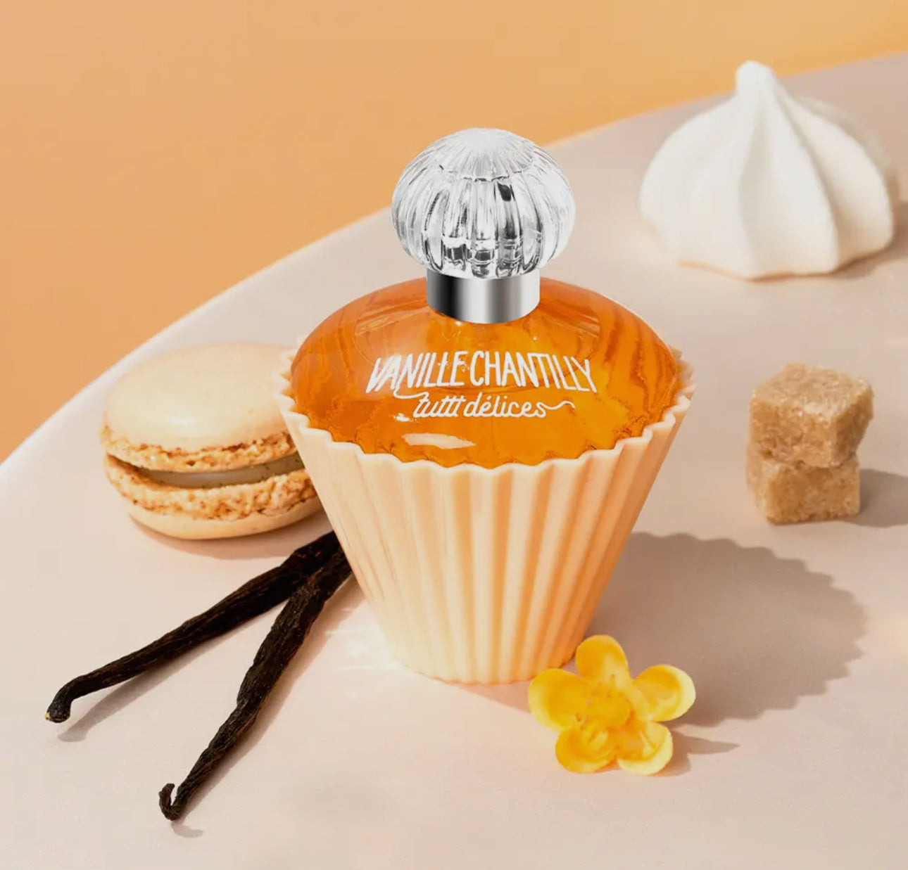 The Vanilla Whipped Cream Cupcake Perfume by Tutti Delices features an amber liquid in a cupcake-shaped bottle with a clear cap, surrounded by a macaron, vanilla beans, yellow flower, sugar cubes, and meringue on a light background.
