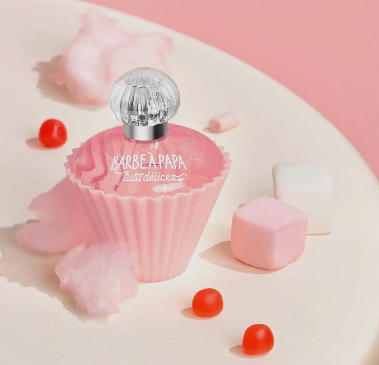 A pink, cupcake-shaped bottle of Cotton Candy Cupcake Perfume by Tutti Delices sits among cotton candy, marshmallows, and red candy balls on a light surface with a pink background.